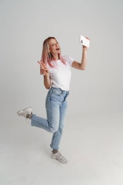 Beyaz arka planda izole edilmiş cep telefonuyla selfie çekerken barış işareti yapan genç bir kadın.