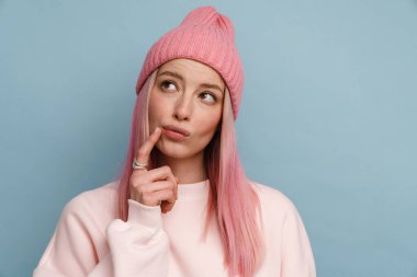 Pembe saçlı genç beyaz kadın, mavi duvarın üzerinden yukarı bakıyor ve düşünüyor.