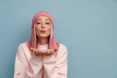 Mavi duvardan izole edilmiş pembe şapkalı genç bir kadın hava öpücüğü veriyor.