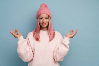 Pembe şapkalı genç kadın gülümsüyor ve mavi duvarın üzerinde meditasyon hareketi yapıyor.