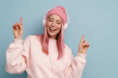 Genç bir kadın, mavi duvarın üzerinden izole edilmiş kulaklıklarla müzik dinlerken parmağıyla yukarı işaret ediyor.