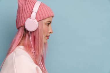 Pembe şapkalı genç bir kadın mavi duvardan izole edilmiş kulaklıklarla müzik dinliyor.