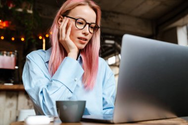 Genç beyaz kadın gözlük takıyor, kafede otururken dizüstü bilgisayarla çalışıyor.