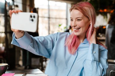 Pembe saçlı genç bir kadın kafede çalışırken cep telefonuyla selfie çekiyor.