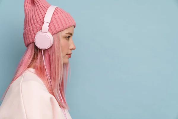戴着粉色帽子的年轻白人妇女带着耳机听着音乐 耳机隔着蓝色的墙壁 — 图库照片
