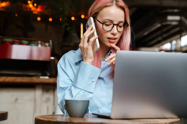 年轻漂亮的女人坐在咖啡店里 一边用笔记本电脑一边用手机说话 — 图库照片