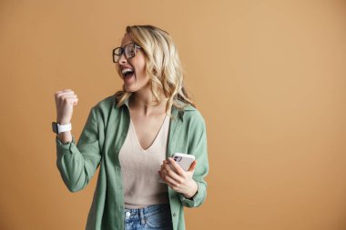 Heyecanlı kadın, bej duvarda izole edilmiş cep telefonu kullanırken kazanan hareketi yapıyor.