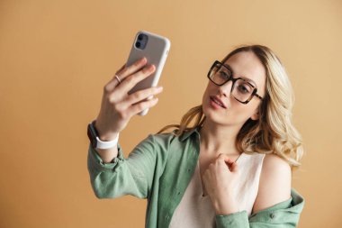 Bej duvarda izole edilmiş cep telefonuyla selfie çeken hoş bir kadın.