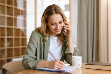 Gülümseyen beyaz sarışın kadın evdeki tahta masada otururken notlar yazıyor.