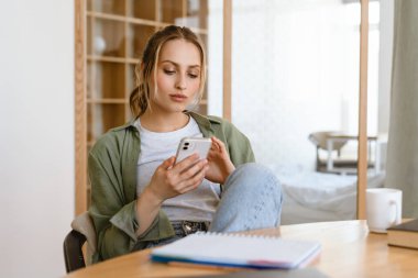 Genç beyaz kadın masada otururken akıllı telefon kullanıyor.