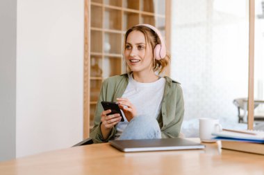 Evde cep telefonu ve kulaklıkla müzik dinlerken gülümseyen genç bir kadın.