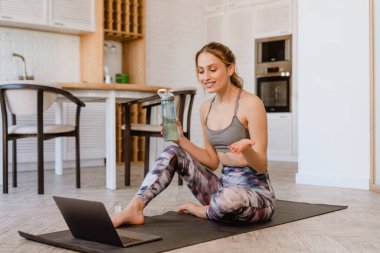 Evde yoga yaparken el kol hareketi yapan ve dizüstü bilgisayar kullanan genç beyaz bir kadın.