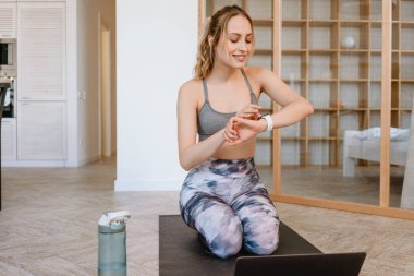 Genç bir kadın evde yoga yaparken akıllı saatlere bakıyor ve laptop kullanıyor.
