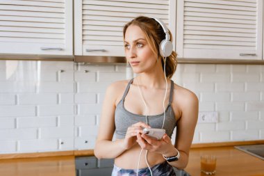 Spor kıyafetli genç beyaz bir kadın meyve suyu içiyor. Evde mutfakta kulaklıkla müzik dinliyor.