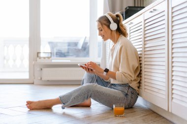 Evde cep telefonu ve kulaklıkla müzik dinlerken gülümseyen genç bir kadın.