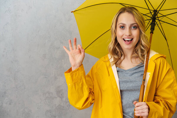 Lovely cheerful young woman wearing waterproof coat holding umbrella isolated over gray background, showing ok