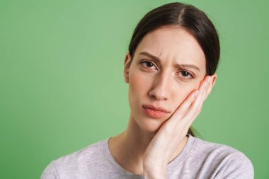 Genç esmer kadın yeşil arka planda tek başına yanağına dokunurken kaşlarını çatıyor.