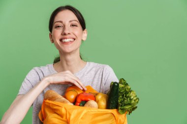 Yeşil arka planda saklanmış sebze dolu tekstil poşetini tutarken gülümseyen genç esmer kadın.