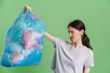 Genç esmer kadın yeşil arka planda izole edilmiş plastik torbalar gösterirken kaşlarını çatıyor.
