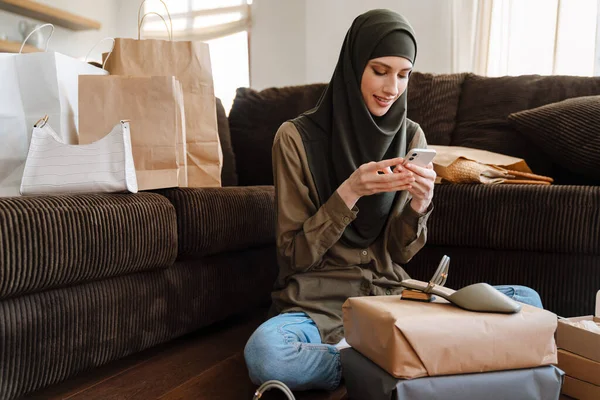 年轻的穆斯林妇女坐在地板上 手里拿着购物袋 一边用手机 — 图库照片