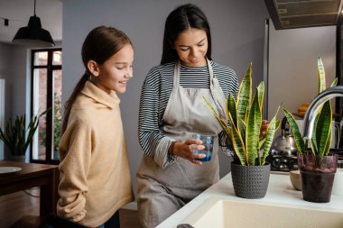 Güzel bir anne ve kız mutfaktaki çiçeklerini sulayıp gülümsüyor.