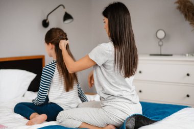 Güzel genç anne, yatakta oturmuş gülümserken küçük tatlı kızının saçlarını tarıyor.