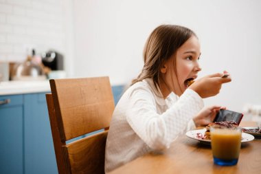 Küçük kız masada oturuyor ve mutfakta krep yiyor.
