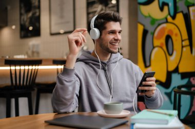 Gülümseyen, kulaklıklı, müzik dinleyen, kafede otururken cep telefonu tutan sıradan bir genç adam.
