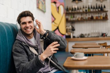 Gülümseyen, kulaklıklı, müzik dinleyen, kafede otururken cep telefonu tutan sıradan bir genç adam.