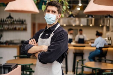 Önlüklü, koruyucu tıbbi maske takan genç bir adam kahve dükkanının kasasında duruyor.