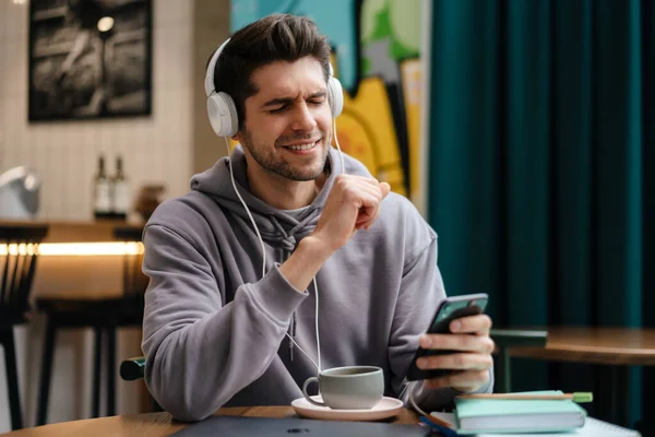 Uśmiechnięty Młodzieniec Słuchawkach Słuchający Muzyki Trzymający Telefon Komórkowy Siedząc Kawiarni — Zdjęcie stockowe