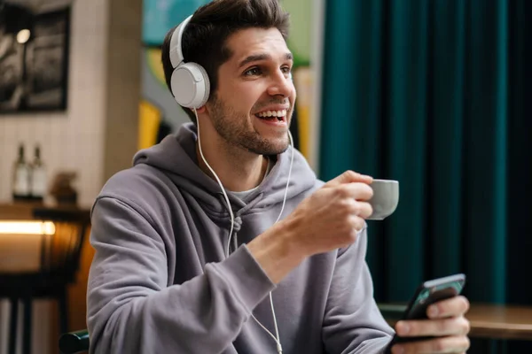 Uśmiechnięty Młodzieniec Słuchawkach Słuchający Muzyki Trzymający Telefon Komórkowy Kawiarni Wewnątrz — Zdjęcie stockowe