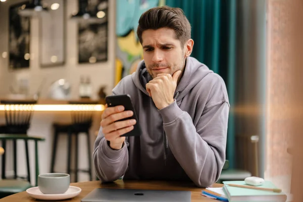 Pensativo Joven Morena Hombre Sosteniendo Teléfono Móvil Mientras Está Sentado — Foto de Stock