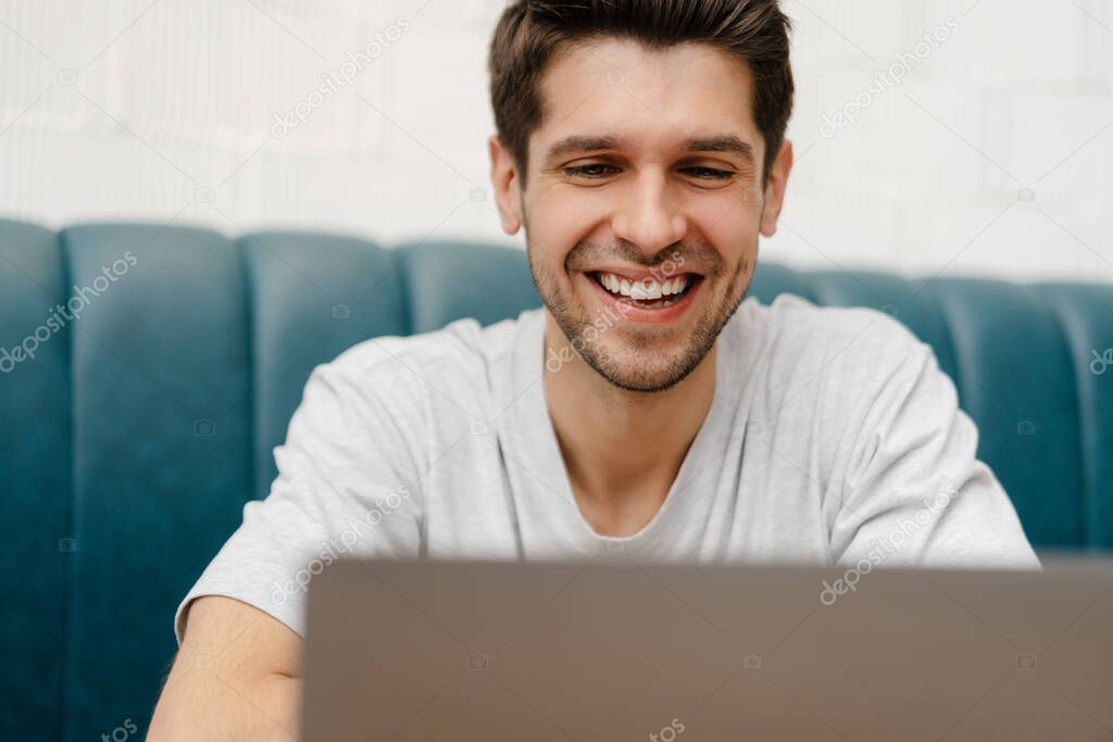 Smiling young man student sitting in cafe indoors with laptop computer, looking at screen