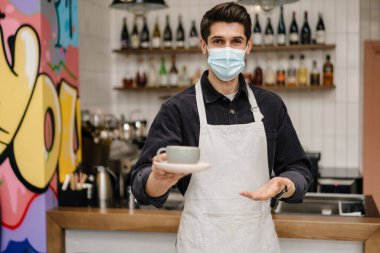 Önlüklü, koruyucu maskeli, elinde kahve tutan esmer, genç bir adam.