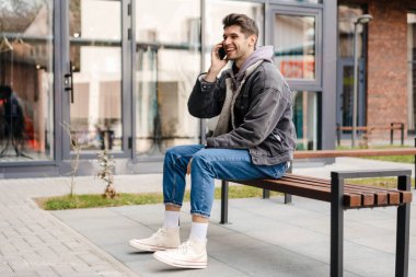 Sıradan kıyafetlerle gülümseyen genç beyaz adam şehrin dışında cep telefonu kullanıyor, bankta oturuyor, konuşuyor.