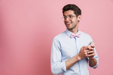 Genç beyaz adam papyon takıyor gülümsüyor ve pembe arka planda cep telefonu kullanıyor.