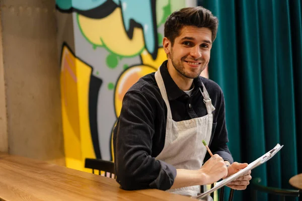 Gülümseyen Genç Garson Kafeteryada Defteriyle Malların Kalitesini Kontrol Ediyor — Stok fotoğraf