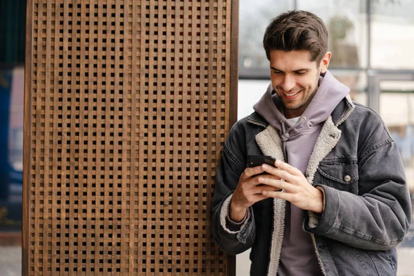 Smiling young white man in casual cothes using mobile phone outdoors in the city, leaning on a wall