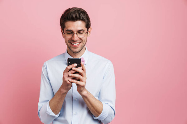 Young white man wearing eyeglasses smiling and using cellphone isolated over pink background