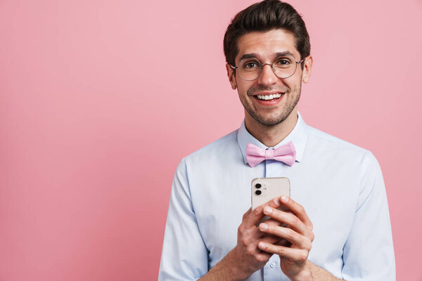 Young white man wearing bow tie smiling and using cellphone isolated over pink background