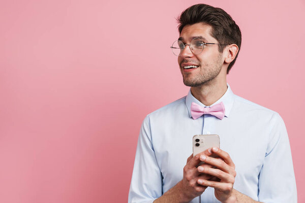 Young white man wearing bow tie smiling and using cellphone isolated over pink background