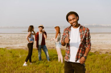 Genç siyahi adam dışarıda arkadaşlarıyla dinlenirken gülümsüyor ve başparmağını gösteriyor.