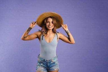Mor arka planda izole edilmiş hasır şapka giyerken gülümseyen genç beyaz kadın.