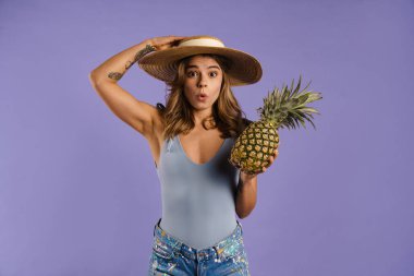 Hasır şapkalı genç beyaz kadın, mor arka planda ananas tutarken şaşkınlığını dile getiriyor.