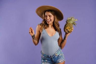 Hasır şapkalı, gülümseyen ve elinde mor arka planda izole edilmiş ananas tutan genç bir kadın.