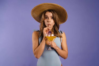 Mavi arka planda duran mayo ve yaz şapkası giyen genç beyaz kadın elinde kokteyl tutuyordu.