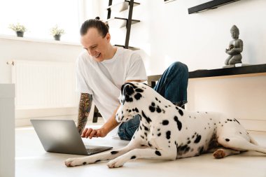 Genç, orta yaşlı bir adam evde köpeğiyle yerde otururken dizüstü bilgisayarda çalışıyor.