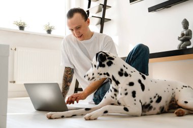 Genç, orta yaşlı bir adam evde köpeğiyle yerde otururken dizüstü bilgisayarda çalışıyor.