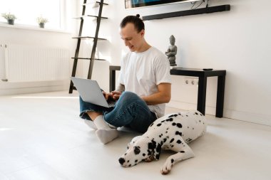 Genç, orta yaşlı bir adam evde köpeğiyle yerde otururken dizüstü bilgisayarda çalışıyor.